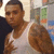 A person in a white tank top with visible tattoos on the right arm, standing against a blurred background with green and blue elements.