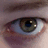 A close-up of a human eye with a brown iris, visible eyelashes, and a reflection of light on the cornea, set against a blurred background.