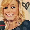 A close-up of a person with blonde hair holding a microphone, wearing a dark polka-dot top, with a black heart symbol in the upper right corner.