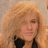 Pixelated image of a person with voluminous light brown hair wearing dark clothing against a neutral background.