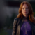 A pixelated image of a person with long reddish-brown hair wearing a dark jacket over a purple top against a blurred dark background.
