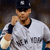 Pixelated image of a person in a white baseball jersey with dark sleeves and cap, holding an object near the face against a blurred background.
