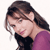 Pixelated image of a person with long brown hair wearing a purple top and dark inner layer, head tilted downward with a slight smile.