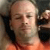 Pixelated image of a person with a bald head wearing an orange garment with a dark red tie, set against a blurred background with dark and light tones.