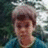 Pixelated static image of a person with light brown hair and dark eyes, wearing a blue garment against a blurred green background.