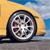 A pixelated car wheel with a silver alloy rim and black tire, positioned on a road with a blurred yellow vehicle and blue sky background.