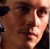 Pixelated close-up of a person's face with brown hair, dark eyes, and light skin, wearing a dark garment against a blurred brown background.