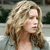 Pixelated image of a person with light brown wavy hair, fair skin, and a neutral expression, cropped to show shoulders and upper chest against a blurred background.