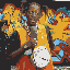 Animated sequence showing a person with dark skin wearing yellow and orange clothing, holding a white drum-like object against a colorful background.