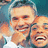 Two people in white shirts smiling and laughing, one with arm around the other, against a blue background.