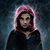A person with pink hooded hair and dark clothing stands against a dark blue background, wearing a black hoodie over a red-collared shirt.