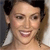 Pixelated close-up of a person with light skin tone, dark hair styled in two buns, wearing large white earrings, and smiling with visible teeth.