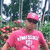 A person in a pink cap and pink shirt with white text stands in a garden with green foliage and orange flowers, captured in a static image with no visible motion.