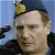 A pixelated close-up of a person wearing a dark blue cap with a yellow emblem, light brown hair, and a dark jacket against a blue background.