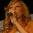 Pixelated static image of a person with long wavy brown hair holding a microphone, set against dark background with light-colored surface below.