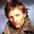 Pixelated close-up of a person with light brown hair and a neutral expression wearing a textured brown jacket against a gradient background.