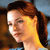 Pixelated close-up of a person with brown hair tied back, wearing a light blue top, against a blurred background with brown and blue tones.