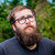 Pixelated portrait of a person with light skin, short brown hair, and a beard, wearing a dark blue shirt against a green foliage background.