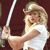 A pixelated image of a person with long blonde hair wearing a white sleeveless top, holding a sword-like object against a dark red background.