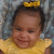 A child in a yellow dress with a ruffled collar and yellow bow headband lies on a patterned surface, smiling with eyes closed in the first frame and eyes open in the second frame.