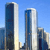 Pixelated image of two tall modern skyscrapers with reflective glass facades against a clear blue sky, with smaller buildings visible in the background.