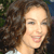 A pixelated close-up of a person with curly brown hair, light skin, and a smiling expression, wearing a white top against a blurred background.