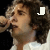 Pixelated close-up of a person with dark curly hair holding a microphone near their mouth, wearing a white top against a black background.
