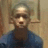 Pixelated static image of a person with light hair wearing a blue top against a muted background with vertical elements.