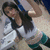 A woman with long dark hair in a white crop top and teal shorts poses against a light background, her right arm raised.