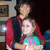 Two people in a pixelated image: one in red and black clothing holding another in a light blue and pink outfit against a dark background with a framed picture.