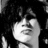 Pixelated grayscale image of a person with dark hair holding a microphone, mouth open as if singing or speaking, against a dark background.