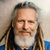 Static pixelated image of a person with long dark hair, light skin, and a white beard, wearing a blue garment against a light background.
