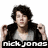 Pixelated image of a person with dark hair wearing a black jacket over a light gray shirt, making fists near the shoulders.