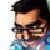 Pixelated close-up of a person with dark hair and glasses, wearing a blue garment against a white background.