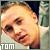 Pixelated close-up of a person's face with light skin tone, visible eyes and mouth, and short hair; text 'TOM' in white block letters at bottom left.