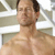 A pixelated close-up of a shirtless person with short brown hair, light skin, and a neutral expression against a blurred background with white and gray tones.
