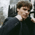 Pixelated image of a person with brown hair wearing dark clothing and white collar, seated against blurred background.