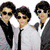 Three figures in formal attire stand together on a white background, with one figure wearing a black tie and sunglasses.