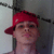 A person wearing a red cap with visible text, standing against a light-colored wall and dark vertical structure.