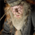 Pixelated image of a figure with light-colored hair, a long white beard, and a dark robe, holding a staff-like object against a dark background.