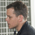 Pixelated profile view of a person with short dark hair wearing a dark jacket over a light shirt against a grid-patterned background.