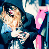 A person with long wavy blonde hair wears a dark hooded jacket and holds a small object while standing near a pink and blue structure.