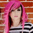 Pixelated image of a person with long pink hair and dark bangs against a brick wall background, wearing a black and white striped top.