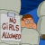Pixelated cartoon icon featuring a character with brown hair and glasses holding a sign that reads 'GIRLS' in large letters above smaller text, set against a light background with blue elements.