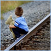 A child in a blue shirt and dark pants crouches on gravel near railroad tracks, holding a light-colored object.