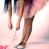A person in a pink tutu and white shoes bends forward, holding a small pink object with both hands against a light pink background.
