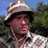 Pixelated image of a person wearing a wide-brimmed hat with light and dark stripes, standing outdoors against trees and blue sky.