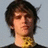 Pixelated static image of a person with dark curly hair, light skin tone, and a neutral expression against a pale blue background.