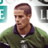 Pixelated static image of a person in a green jacket with dark collar and white shirt, set against light background with partial teal and gray shapes.
