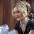 Static pixelated image of a person with blonde hair in an updo wearing a dark top and turquoise necklace, seated at a table with a white object under dim lighting.
